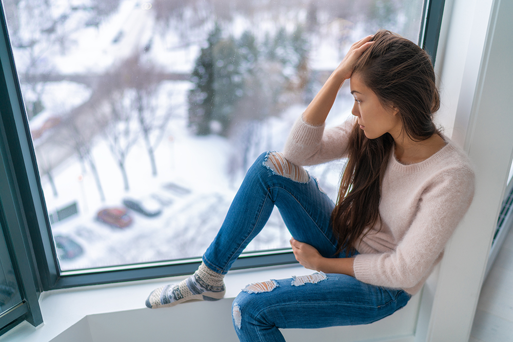 Winter depressed sad girl lonely by home window looking at cold weather upset unhappy. .